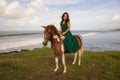 Happy woman riding horse near the ocean. Outdoor activities. Asia woman wearing long green dress. Traveling concept. Cloudy sky. Royalty Free Stock Photo