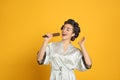 Happy young woman in silk bathrobe with hair curlers singing into hairbrush on orange background Royalty Free Stock Photo