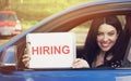 Happy woman sitting inside car showing white card with hiring sign message