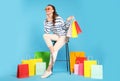 Happy woman in stylish sunglasses holding colorful shopping bags on stool against light blue background Royalty Free Stock Photo