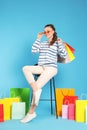 Happy woman in stylish sunglasses holding colorful shopping bags on stool against light blue background Royalty Free Stock Photo