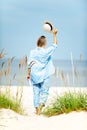 Happy woman with summer hat holding book walking on seaside sand. Outdoors vertical colored image. View from backside Royalty Free Stock Photo