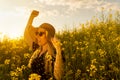 Happy woman in sun hat and glasses dancing and enjoying summer raising arms in yellow flowers field at sunset Royalty Free Stock Photo