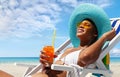 Happy woman is sunbathing on a beach deck chair, wearing sun hat and sunglasses, drinking a orange juice on a sunny day by the Royalty Free Stock Photo