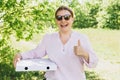 Happy Woman in sunglasses holding a box with pizza and showing thumbs up on nature background. Fast pizza delivery. Mock Royalty Free Stock Photo