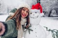 Happy woman takes selfie by snowman in Santa hat outdoors in snowy winter park. Christmas festive season Royalty Free Stock Photo