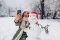 Happy woman takes selfie by snowman in Santa hat outdoors in snowy winter park. Christmas festive season Royalty Free Stock Photo