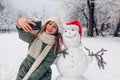 Happy woman takes selfie by snowman in Santa hat outdoors in snowy winter park. Christmas festive season Royalty Free Stock Photo