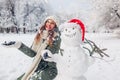 Happy woman tossing snow up by snowman in Santa hat outdoors in snowy winter park. Christmas festive season Royalty Free Stock Photo