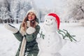 Happy woman tossing snow up by snowman in Santa hat outdoors in snowy winter park. Christmas festive season Royalty Free Stock Photo