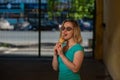 A happy woman in a turquoise dress stands in the yard and eats a waffle cone on a warm summer day. Beautiful blonde in sunglasses