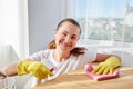 Happy woman in white shirt and yellow protective rubber gloves cleaning at home and wiping dust on wood table. Royalty Free Stock Photo