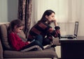 Happy woman working at home on computer and cute kid studing in mobile phone. Family sitting on sofa together near each other. Royalty Free Stock Photo