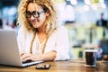 Happy woman working on laptop at coffee shop. Woman in eyeglasses and beads necklace using laptop on desk with mobile phone and Royalty Free Stock Photo