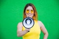 Happy woman in a yellow dress glasses and earrings on a green background shouting into a megaphone. Portrait of a girl Royalty Free Stock Photo