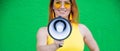 Happy woman in a yellow dress glasses and earrings on a green background shouting into a megaphone. Portrait of a girl Royalty Free Stock Photo