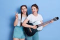 Happy women friends with guitar and showing v sign, keeping mouths opened, wearing white t shirt and jeans shorts, posing isolated Royalty Free Stock Photo