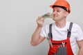 Happy worker in red uniform, protective hard hat holding bundle of dollars, sniffing of cash money on white background. Male Royalty Free Stock Photo