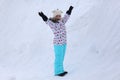 Happy 8 years old Caucasian girl in colored overalls is standing on a mountain with lifted hands on snow outside in winter