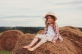 Happy 7 years old child girl in country style plaid shirt and hat relaxing on summer field with hay stacks