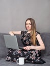 Happy young adorable woman in long dress sitting on sofa with laptop and big white mug at home