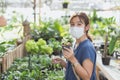 Happy young adult Asian woman with medical face mask holding pot, little tree pot, pot of flowers or plants at greenhouse or Royalty Free Stock Photo