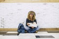 Happy young adult traveler woman sitting on the floor reading a book. Travel female people lifstyle. Curly hair and trendy hat Royalty Free Stock Photo