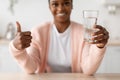 Happy young african american lady hold glass of clean water and show thumb up in minimalist kitchen