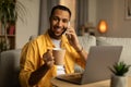 Happy young African American man with coffee speaking on cellphone, using laptop pc at home office Royalty Free Stock Photo