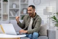 Happy young african american man using laptop on couch at home. Celebrates, rejoices, received good news, watches online Royalty Free Stock Photo