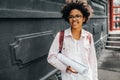 Happy young african american woman smiling with backpack and paper roll outdoors. Cheerful afro student female wearing eyeglasses Royalty Free Stock Photo