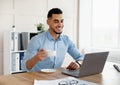 Happy young Arab man drinkng coffee while using laptop at modern office