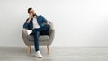 Happy young Arab man talking on smartphone, sitting in armchair against white studio wall, free space