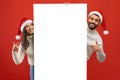 Happy young arab man and woman in Santa hats posing by empty advertising board, showing xmas offer, banner