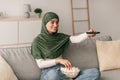 Happy young Arab woman with remote control sitting on couch, watching TV and eating popcorn at home Royalty Free Stock Photo