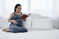 Happy young Asia pregnant woman holding and reading red book and sitting on white bed Royalty Free Stock Photo