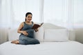 Happy young Asia pregnant woman holding and reading red book and sitting on white bed Royalty Free Stock Photo