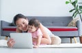 Happy young Asian beautiful mother sitting on floor, working with tablet while holding her cute little Caucasian 7 months newborn