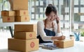 Happy young Asian business owner woman prepare parcel box and check online orders of product for deliver to customer on smartphone Royalty Free Stock Photo