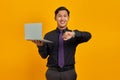 Happy young Asian businessman looking at wristwatch and holding laptop isolated on yellow background Royalty Free Stock Photo