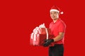 Happy young Asian delivery man in red uniform, Christmas hat carry boxes of presents in hands on red background during Christmas f