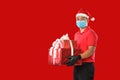 Happy young Asian delivery man in red uniform, medical face mask, protective gloves, Christmas hat carry boxes of presents in hand