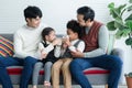 Happy young Asian gay couple with diverse adopted children African and Caucasian holding glass of milk sitting on sofa at home. Royalty Free Stock Photo