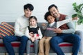 Happy young Asian gay couple with diverse adopted children African and Caucasian smiling sitting on sofa at home.