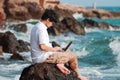 Happy young Asian man with computer laptop sitting on the rock of seashore in summer time. Summer vacations and lifestyle concept