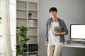 A happy young Asian man in a flannel shirt stands in his modern office with books in his hand