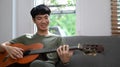 Happy young asian man playing acoustic guitar, spending leisure time in cozy living room Royalty Free Stock Photo