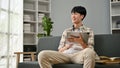 A happy young Asian man using his digital tablet while relaxing on a sofa in the living room Royalty Free Stock Photo