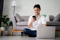 Happy young asian man using mobile phone while sitting at a couch at home with laptop computer Royalty Free Stock Photo