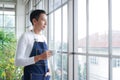 Happy young Asian man wearing apron smiling and holding cup of coffee looking out of window after take care of trees in greenhouse Royalty Free Stock Photo